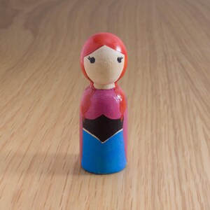 May include: A wooden peg doll with a painted design. The doll has a red head, a light skin tone face, and is wearing a pink, black, and blue dress. The doll is standing on a wooden surface.