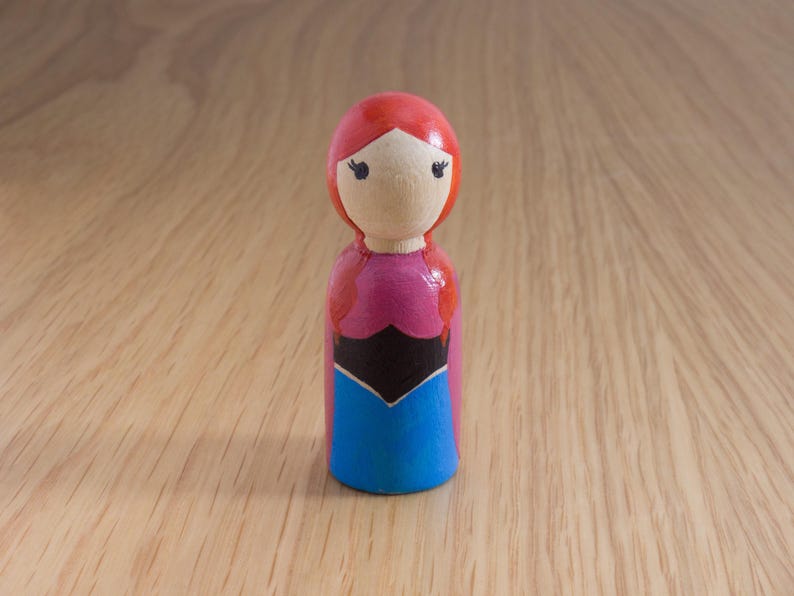May include: A wooden peg doll with a painted design. The doll has a red head, a light skin tone face, and is wearing a pink, black, and blue dress. The doll is standing on a wooden surface.