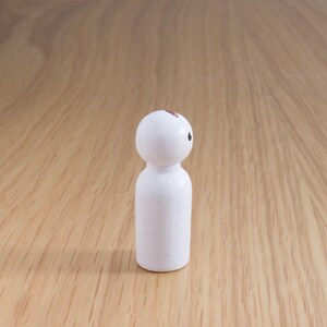 May include: A small, white wooden peg doll with a rounded head and cylindrical body. The doll has a single black eye and a small brown mark on its head. The doll is standing on a light brown wooden surface.