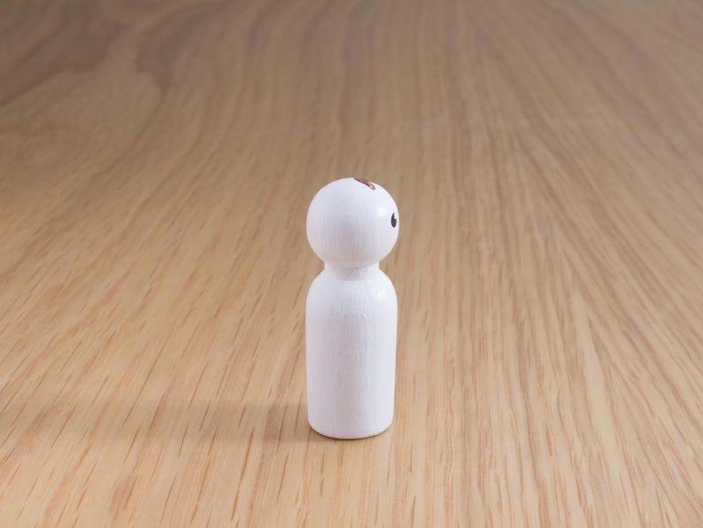 May include: A small, white wooden peg doll with a rounded head and cylindrical body. The doll has a single black eye and a small brown mark on its head. The doll is standing on a light brown wooden surface.
