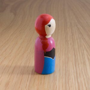 May include: A wooden peg doll with a red head and hair, a light-colored face, a pink torso, a black accent, and a blue skirt. The doll is standing on a light brown wooden surface.