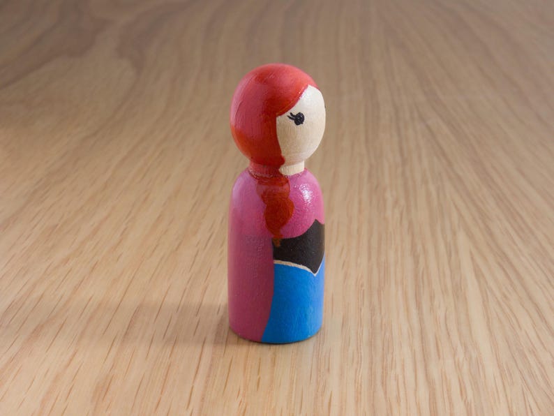May include: A wooden peg doll with a red head and hair, a light-colored face, a pink torso, a black accent, and a blue skirt. The doll is standing on a light brown wooden surface.