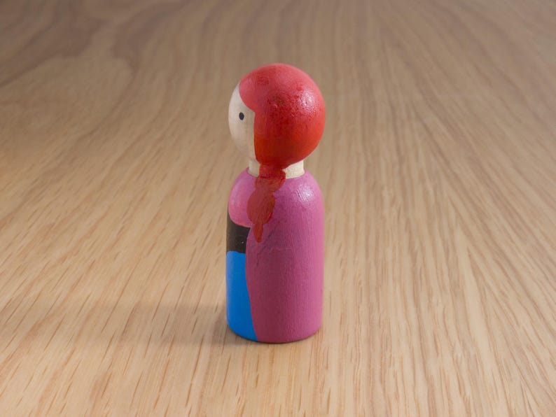 May include: A wooden peg doll with a red head and a pink body. The doll has a blue section on the bottom and a black stripe. The doll is painted with a simple design and stands on a wooden surface.