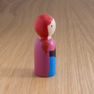 May include: A painted wooden peg doll with a red head and hair, a white face, and a pink body. The doll has a black and blue section on the side. The doll is standing on a wooden surface.
