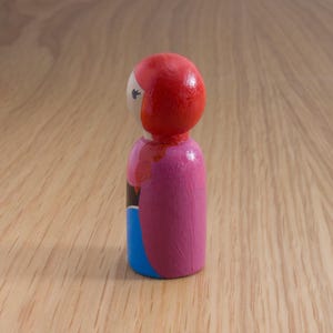 May include: A hand-painted wooden peg doll with a red head, pink body, and blue skirt. The doll has a simple design with black and white details. The doll is standing on a wooden surface.