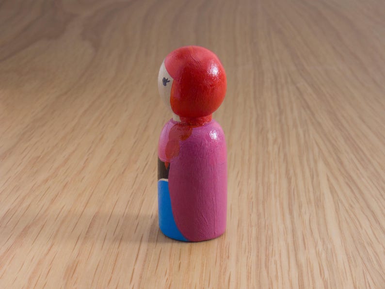 May include: A hand-painted wooden peg doll with a red head, pink body, and blue skirt. The doll has a simple design with black and white details. The doll is standing on a wooden surface.