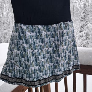 May include: A black ribbed waistband skirt with a forest print. The skirt features a pattern of green and blue trees on a white background. The hemline is trimmed with a geometric black and gray design. The skirt is hanging on a clothes hanger.