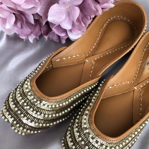 Puede incluir: Un par de zapatos planos adornados, con un interior de cuero marr&oacute;n. Las partes superiores est&aacute;n cubiertas con filas de lentejuelas doradas y peque&ntilde;as perlas blancas. Los zapatos se exhiben sobre una superficie de sat&eacute;n plateado, con un ramo de flores moradas en el fondo.