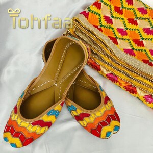 K&ouml;nnte beinhalten: Ein Paar bunte, bestickte flache Schuhe mit einem geometrischen Muster in Gelb, Rot, Orange und Blau. Die Schuhe sind aus Leder und haben eine braune Ledersohle.