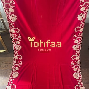 Puede incluir: Un textil rojo vibrante con intrincados bordados florales en los bordes. El diseño presenta un patrón repetitivo de flores y vides en un color contrastante. El texto "Tohtaa London" se muestra en dorado.