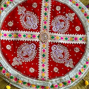 Puede incluir: Una bandeja decorativa redonda roja y ornamentada con intrincados bordados y adornos. La bandeja presenta un diseño central con cuatro formas de tipo paisley, adornadas con cuentas blancas y detalles dorados. El borde está decorado con patrones geométricos coloridos y borlas.