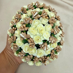 Pode incluir: Um arranjo floral redondo com rosas artificiais na cor creme intercaladas com bagas decorativas em tom dourado metálico e pequenos detalhes dourados texturizados. O conjunto de flores é densamente compacto com bases de folhas verdes.