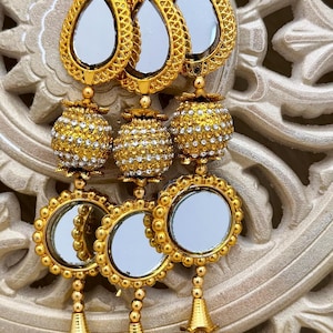 May include: Gold-toned jewelry with mirrored accents and intricate detailing. The design features teardrop-shaped elements, beaded spheres, and circular mirrors, all suspended from a gold chain and finished with brown tassels.