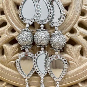 May include: Silver-toned decorative ornaments with mirrored teardrop and round elements, adorned with sparkling embellishments and tassels. The ornaments are suspended against a carved, golden-hued background, creating a striking contrast. The design suggests a festive or ornamental purpose.