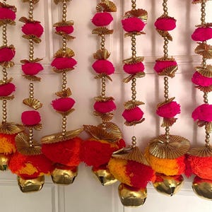 Puede incluir: Guirnalda decorativa con hileras de pompones rosas, cuentas doradas y adornos en forma de campana. La guirnalda tiene detalles dorados en forma de abanico y borlas naranjas y amarillas. Las campanas son doradas.