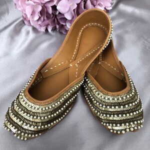 Puede incluir: Un par de bailarinas color canela adornadas con lentejuelas doradas y cuentas blancas con forma de perla. Los zapatos tienen la punta afilada y est&aacute;n decorados con filas de lentejuelas y cuentas. Un ramo de flores moradas est&aacute; en el fondo.