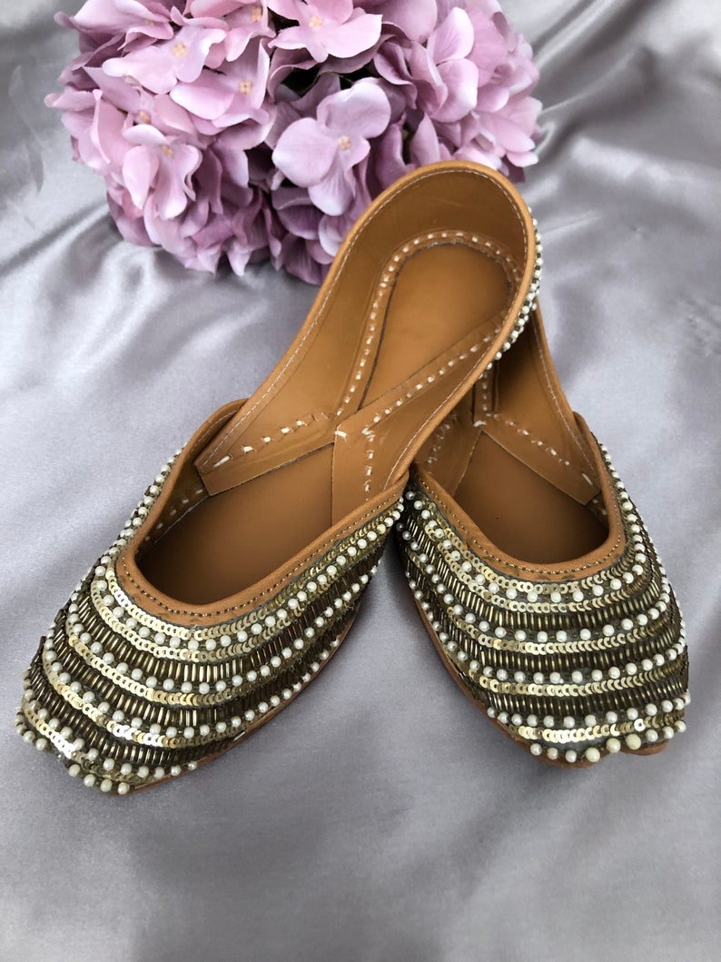Puede incluir: Un par de bailarinas color canela adornadas con lentejuelas doradas y cuentas blancas con forma de perla. Los zapatos tienen la punta afilada y est&aacute;n decorados con filas de lentejuelas y cuentas. Un ramo de flores moradas est&aacute; en el fondo.