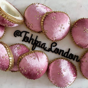 Peut inclure: Plusieurs objets décoratifs en velours rose, certains ronds et d'autres coupés en deux, avec des bordures dorées et des ornements dorés épars. Les objets sont disposés sur une surface blanche, avec le texte "@Tohfaa London" en noir.