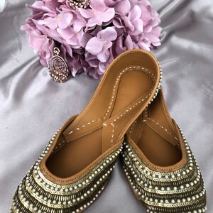 Puede incluir: Un par de zapatos sin cordones de cuero color canela con intrincados adornos de cuentas y lentejuelas. Los zapatos tienen una punta redondeada y est&aacute;n adornados con filas de lentejuelas doradas y cuentas blancas. Un collar con colgante a juego y un ramo de flores rosas en el fondo.