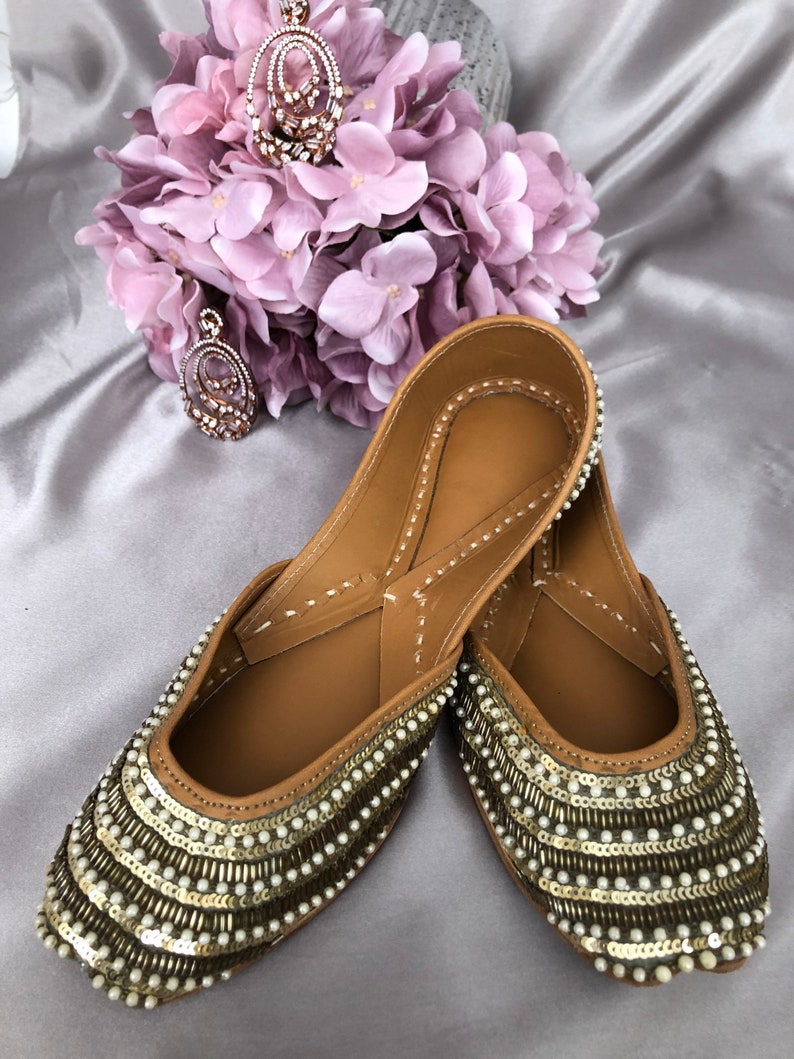 Puede incluir: Un par de zapatos sin cordones de cuero color canela con intrincados adornos de cuentas y lentejuelas. Los zapatos tienen una punta redondeada y est&aacute;n adornados con filas de lentejuelas doradas y cuentas blancas. Un collar con colgante a juego y un ramo de flores rosas en el fondo.