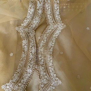 Könnte beinhalten: Nahaufnahme eines goldfarbenen Stoffes mit aufwendiger Stickerei. Das Design zeigt florale Muster und Perlenarbeiten entlang der Kanten, wodurch ein zartes und verziertes Aussehen entsteht. Der Stoff hat eine transparente, geschichtete Textur.