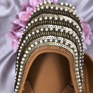 Puede incluir: Primer plano de un zapato de cuero marr&oacute;n con adornos intrincados. La parte superior del zapato est&aacute; adornada con filas de lentejuelas doradas, cuentas blancas y cuentas marrones. El zapato tiene una suela de cuero marr&oacute;n claro y un dise&ntilde;o decorativo.
