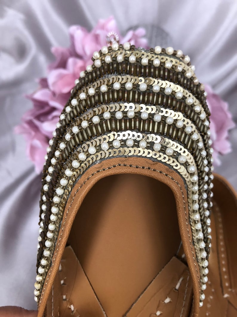 Puede incluir: Primer plano de un zapato de cuero marr&oacute;n con adornos intrincados. La parte superior del zapato est&aacute; adornada con filas de lentejuelas doradas, cuentas blancas y cuentas marrones. El zapato tiene una suela de cuero marr&oacute;n claro y un dise&ntilde;o decorativo.