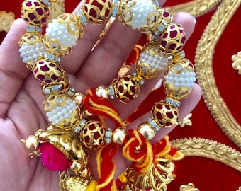 May include: A gold and pearl beaded bracelet with red accents and a decorative tassel. The bracelet features gold filigree beads with red centers, interspersed with white pearl beads. The tassel includes bells and colorful accents.