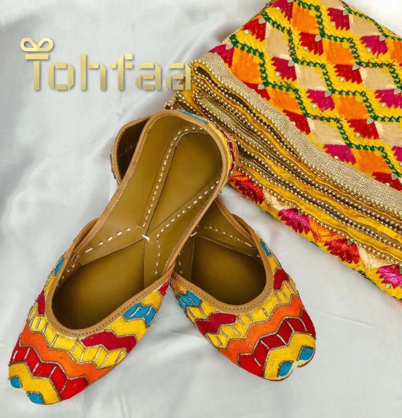K&ouml;nnte beinhalten: Ein Paar bunte, bestickte flache Schuhe mit einem geometrischen Muster in Gelb, Rot, Orange und Blau. Die Schuhe sind aus Leder und haben eine braune Ledersohle.