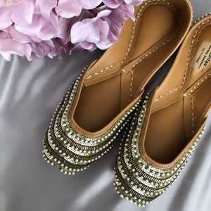 Puede incluir: Un par de bailarinas color beige adornadas con intrincados detalles de cuentas y lentejuelas. Los zapatos tienen una punta redondeada y est&aacute;n adornados con hileras de peque&ntilde;as cuentas blancas y lentejuelas met&aacute;licas. Un racimo de flores de color p&uacute;rpura claro est&aacute; en el fondo.