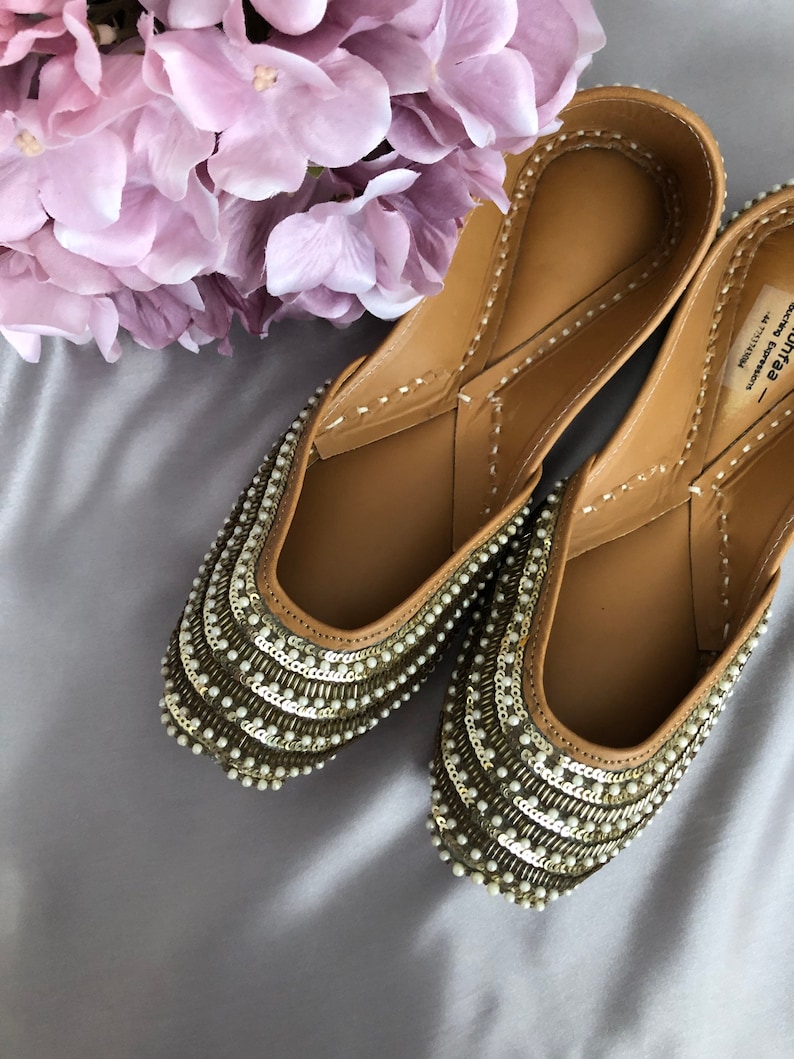 Puede incluir: Un par de bailarinas color beige adornadas con intrincados detalles de cuentas y lentejuelas. Los zapatos tienen una punta redondeada y est&aacute;n adornados con hileras de peque&ntilde;as cuentas blancas y lentejuelas met&aacute;licas. Un racimo de flores de color p&uacute;rpura claro est&aacute; en el fondo.