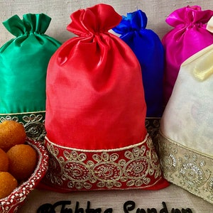 Puede incluir: Cuatro bolsas de cordón de satén de colores con ribetes dorados y adornos de cuentas. Las bolsas están dispuestas en fila sobre una superficie beige. El texto "@Tahftaa London" es visible en primer plano.