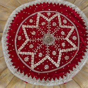 Puede incluir: Una pieza decorativa redonda de terciopelo rojo con intrincados bordados y adornos plateados. El diseño presenta un motivo floral central y un patrón en forma de estrella, rodeado por un borde de cuentas plateadas. El fondo es una tela dorada plisada.