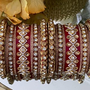 Puede incluir: Un conjunto de pulseras rojas y doradas adornadas con piedras preciosas claras y ámbar y detalles dorados. El diseño presenta patrones de diamantes y florales. Joyería tradicional.