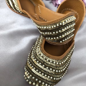 Puede incluir: Un par de bailarinas adornadas con un interior de cuero marr&oacute;n. El exterior est&aacute; decorado con filas de lentejuelas doradas y peque&ntilde;as cuentas blancas. Los zapatos tienen una punta afilada y un tac&oacute;n bajo, adecuados para uso casual o formal.