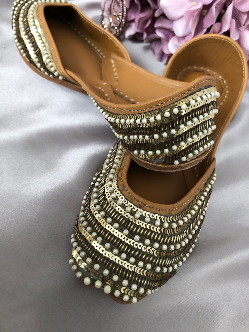 Puede incluir: Un par de bailarinas adornadas con un interior de cuero marr&oacute;n. El exterior est&aacute; decorado con filas de lentejuelas doradas y peque&ntilde;as cuentas blancas. Los zapatos tienen una punta afilada y un tac&oacute;n bajo, adecuados para uso casual o formal.