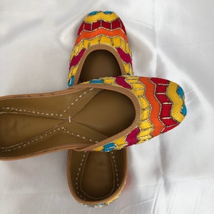 K&ouml;nnte beinhalten: Ein Paar bunte, bestickte flache Schuhe mit einem gelben, orangefarbenen, roten und blauen Muster. Die Schuhe sind aus Leder und haben eine spitze Zehenpartie.