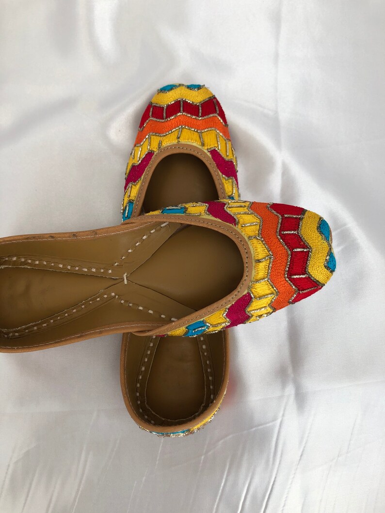 K&ouml;nnte beinhalten: Ein Paar bunte, bestickte flache Schuhe mit einem gelben, orangefarbenen, roten und blauen Muster. Die Schuhe sind aus Leder und haben eine spitze Zehenpartie.