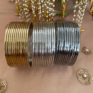 May include: Three stacks of metal bangles in gold, silver, and gunmetal gray. The bangles are round and have a ribbed texture. The background includes gold-toned jewelry with pearls and leaf accents, and decorative gold-toned embellishments.