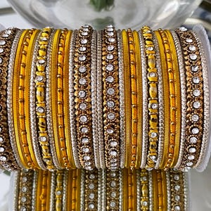 Op de afbeelding: Een stapel gele en gouden armbanden met ingewikkelde details. De armbanden hebben een mix van texturen, waaronder gladde gele banden, kralen secties en banden versierd met heldere, sprankelende stenen. Het algehele ontwerp is sierlijk en opvallend.