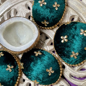May include: An overhead view presents a halved coconut alongside several emerald green velvet coasters. The coasters feature gold trim and sparkling floral details. The coconut's interior is white, and the arrangement rests on a decorative, carved surface.