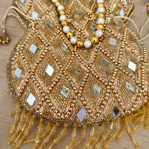 May include: A gold beaded pouch with a drawstring closure. The pouch is covered in gold beads, sequins, and small mirrored squares. A beaded pearl necklace is draped over the top. The bottom of the pouch has dangling gold beaded fringe.