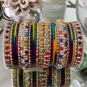 May include: A collection of vibrant bangles with gold detailing and sparkling rhinestones. The bangles feature a mix of solid colours and embellished designs, reflecting light. The image shows a variety of colours, including red, blue, yellow, and green.