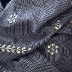 May include: Close-up of a draped, textured fabric in a muted grey-blue hue. The fabric features vertical stripes and is embellished with mirrored floral and leaf-shaped accents. The overall aesthetic suggests a garment or textile.