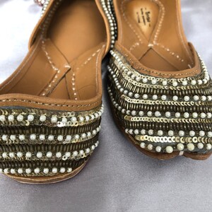 Puede incluir: Un par de zapatos planos adornados. Los zapatos son de color beige con filas de lentejuelas doradas y peque&ntilde;as cuentas blancas. Las suelas son de color marr&oacute;n claro. Los zapatos est&aacute;n decorados con un patr&oacute;n de lentejuelas y cuentas.