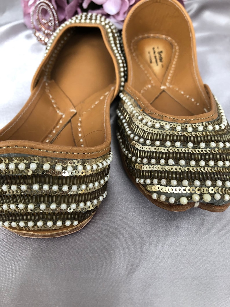 Puede incluir: Un par de zapatos planos adornados. Los zapatos son de color beige con filas de lentejuelas doradas y peque&ntilde;as cuentas blancas. Las suelas son de color marr&oacute;n claro. Los zapatos est&aacute;n decorados con un patr&oacute;n de lentejuelas y cuentas.