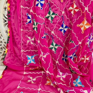 Peut inclure: Un textile rose vif avec des broderies complexes. Le motif présente des motifs géométriques bleus, verts, oranges et blancs, reliés par de fins fils argentés. Les bords sont ornés de coutures décoratives colorées, créant un aspect riche et détaillé.