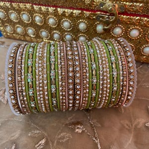 Puede incluir: Un conjunto de pulseras verdes, doradas y blancas con detalles intrincados. Las pulseras están adornadas con pequeñas perlas blancas y gemas transparentes. En el fondo, una caja decorativa dorada con detalles de perlas.