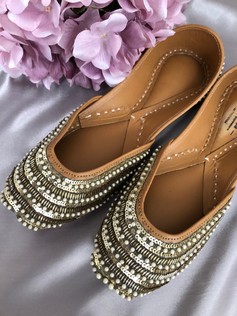 Puede incluir: Un par de zapatos planos adornados, con un interior de cuero marr&oacute;n. Las partes superiores est&aacute;n cubiertas con filas de lentejuelas doradas y peque&ntilde;as perlas blancas. Los zapatos se exhiben sobre una superficie de sat&eacute;n plateado, con un ramo de flores moradas en el fondo.