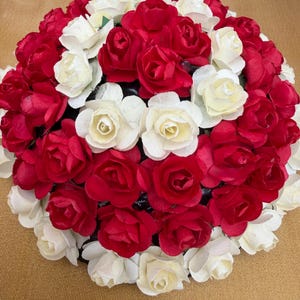 May include: A spherical floral arrangement featuring a dense cluster of artificial roses in alternating shades of deep red and off-white. The roses are tightly packed together, creating a rounded, dome-like shape with layered petals.